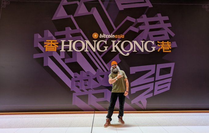A bearded man wearing an orange cap and olive shirt poses playfully in front of a large purple "Bitcoin Asia" backdrop featuring "Hong Kong" prominently displayed in English and Chinese characters, capturing a moment from Day 2 of the Bitcoin Asia conference in Hong Kong.