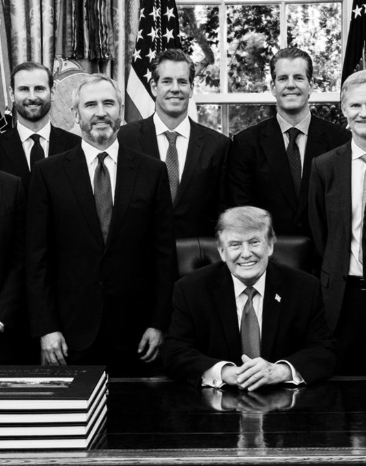 The black-and-white photo shows President Donald Trump seated at the Resolute Desk in the Oval Office, smiling, surrounded by a group of men in business suits standing behind him, with American flags and the presidential seal visible in the background. The image appears to be from an official White House signing or meeting event, though no specific text or names are clearly visible in the photo itself.