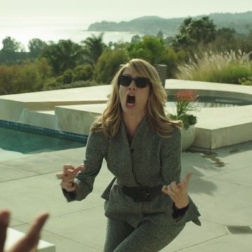 A blonde woman wearing sunglasses and a grey blazer kneels outdoors by a pool with her mouth open in an expressive gesture.
