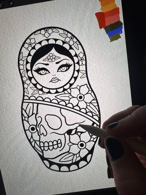 An artist using a stylus on a tablet to color a black and white line art illustration of a Russian nesting doll (matryoshka) featuring a woman with flowers and a skull design, with a rainbow pride flag visible in the corner.