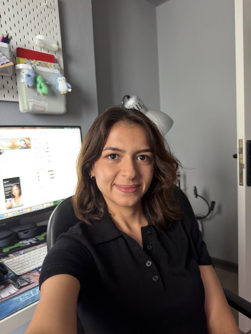 A selfie of a woman with shoulder-length brown hair wearing a black polo shirt, sitting in front of a computer monitor in a home office with a pegboard and small plush toys on the wall.