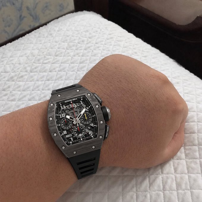 A person's wrist displays a luxury Richard Mille chronograph watch with a black carbon-fiber tonneau case, skeletonized dial, and black rubber strap, resting against a white quilted surface, serving as visual "proof of profits" to flex the wearer's successful taste in high-end horology.