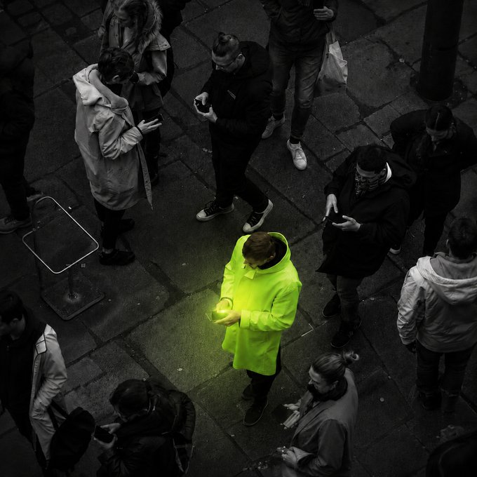 A lone figure in a vivid neon-green jacket stands out dramatically among a crowd of muted, black-clad pedestrians on a dimly lit cobblestone plaza, their phone screen casting a matching green glow on the ground. The image visually mirrors the "bullish thesis, defensive sizing" sentiment—the bright outlier symbolizing selective optimism amid a largely cautious, somber crowd.