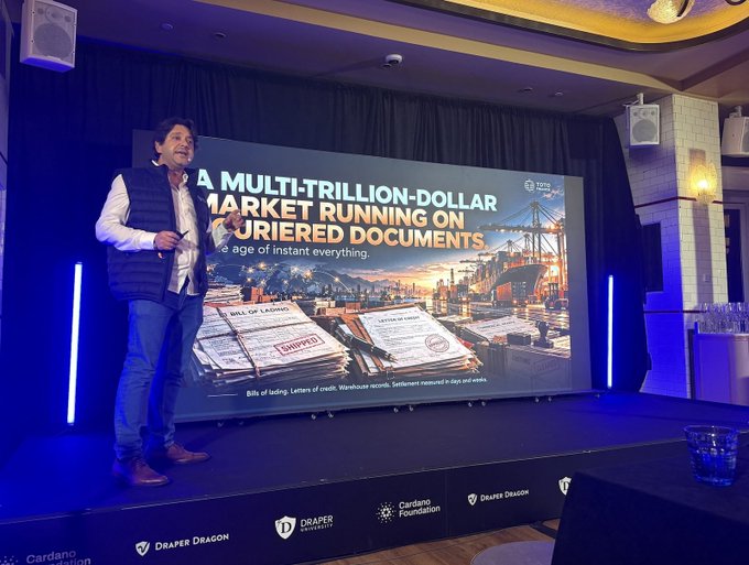 A woman giving a presentation on a stage in front of a large screen titled 'A MULTI-TRILLION-DOLLAR MARKET RUNNING ON BURIED DOCUMENTS,' with logos for Draper Dragon and Cardano Foundation displayed on the stage.