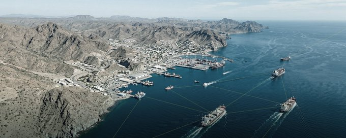 Aerial view of a coastal port city nestled among rugged, arid mountains, with several ships and boats visible in the blue harbor waters.