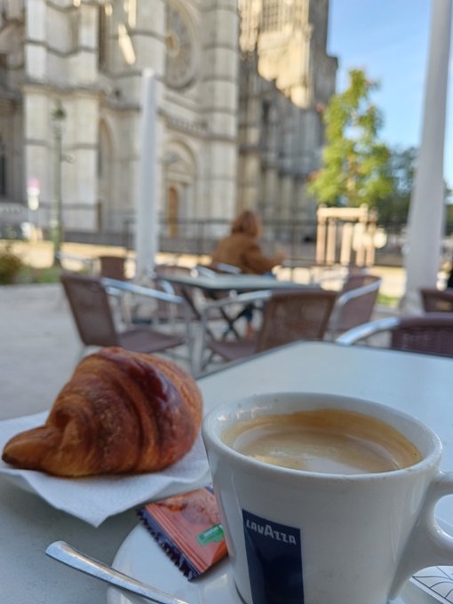 A Lavazza espresso cup and a croissant sit on an outdoor café table, with a Gothic-style cathedral visible in the background.