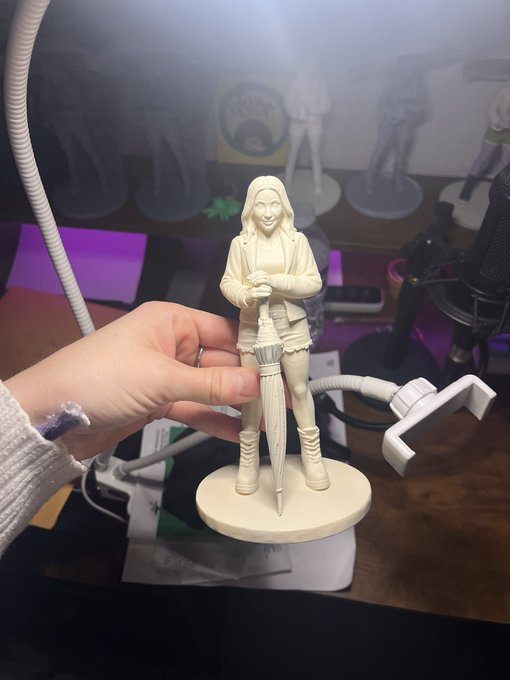 A hand holding an unpainted resin figurine of a female character holding an umbrella, with other similar figurines visible on a shelf in the background near a desk lamp.