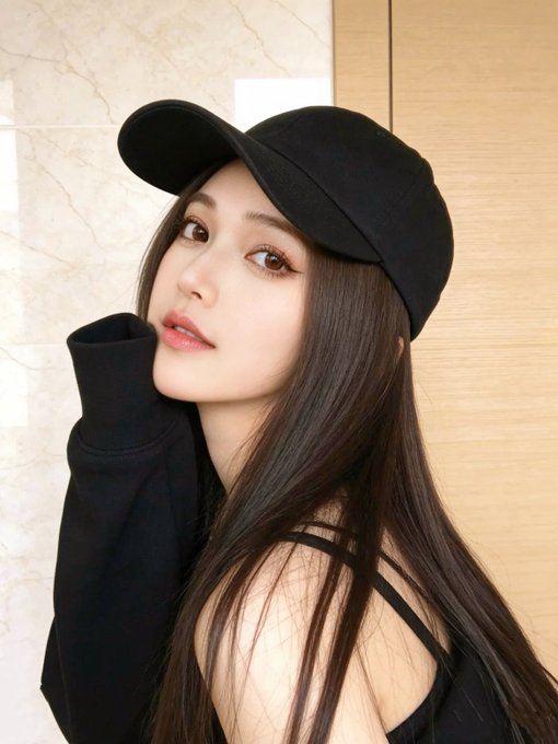 A close-up portrait of a young woman with long dark hair wearing a black cap and a black top with cut-out shoulder straps.