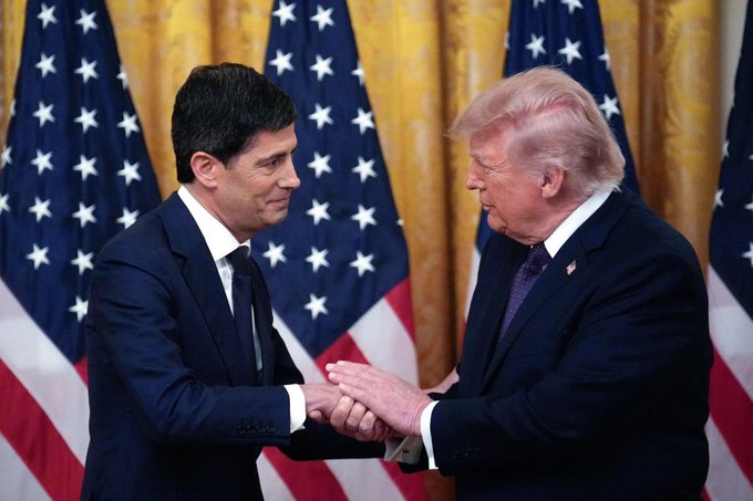 Two men in dark suits shake hands in front of American flags and gold curtains, with Donald Trump on the right and another official on the left. The image visually accompanies Trump's call for the Federal Reserve to lower interest rates to 1%, reflecting his push for monetary policy changes to support his economic agenda.