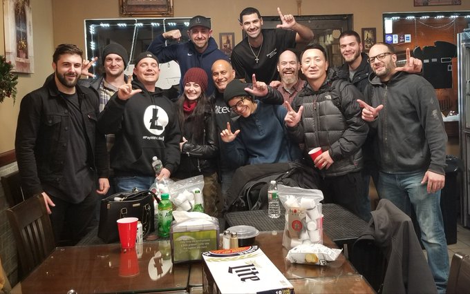 A group photo of around a dozen people posing together indoors, smiling and making hand gestures, with drinks and snacks on a table in front of them.