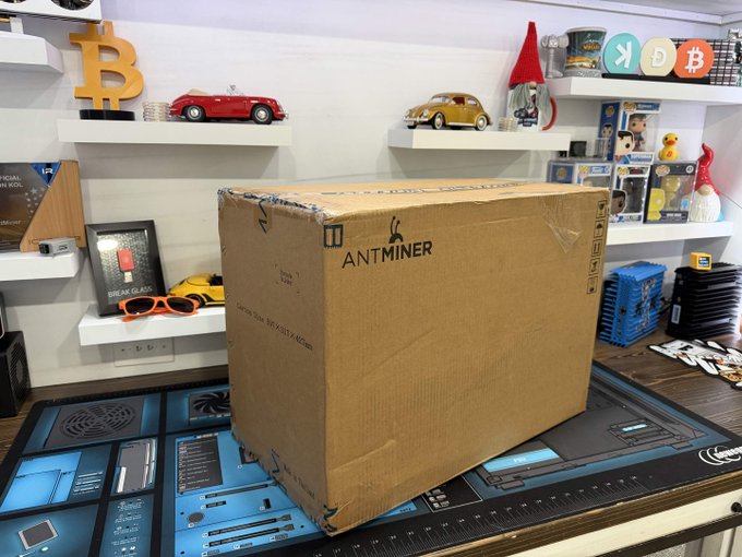 urns, figurines, and a blue and white sign with "ANTMINER" written on it are displayed on shelves above a desk with a black mouse pad.