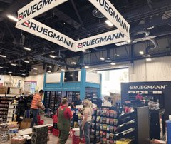 A photo of a BRUEGMANN retail/trade show booth at an indoor convention, with branded hanging banners and people browsing shoe displays below.
