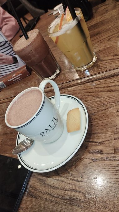 A wooden table displays a tall mug of frothy hot chocolate branded 'PAUL' with a biscuit on the saucer, alongside a glass of iced orange drink and a dark blended beverage with straws.