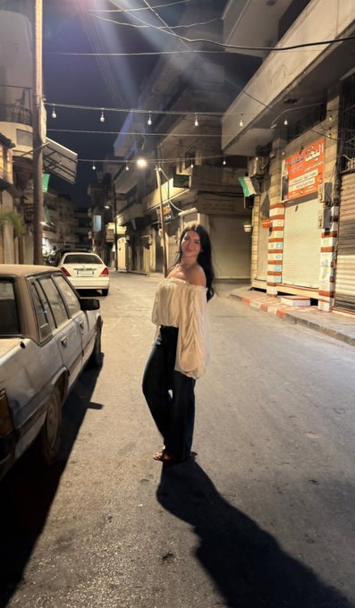 A nighttime photograph of a woman in a white off-shoulder top and dark jeans posing on a street lit by string lights, with closed shops featuring Arabic signage visible in the background.