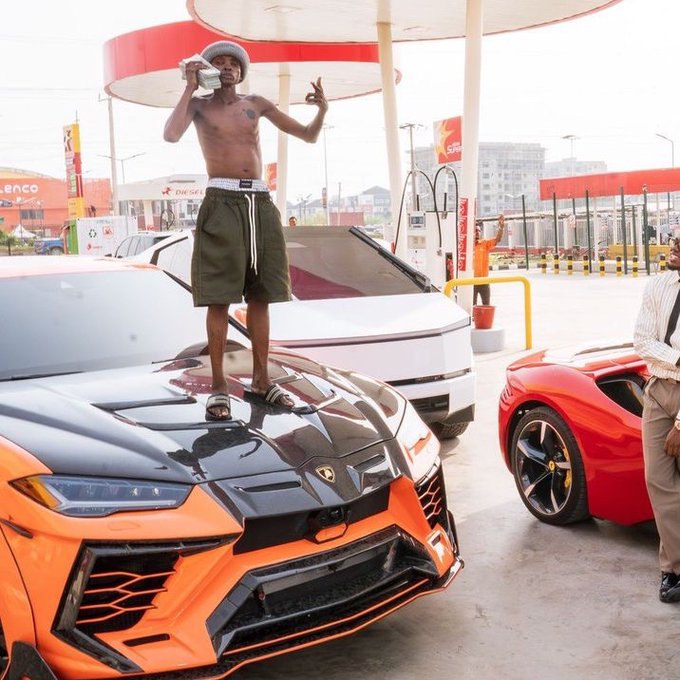 A shirtless man wearing a knit cap and green shorts stands on the hood of an orange and black Lamborghini at a gas station, while a red Ferrari is parked nearby.