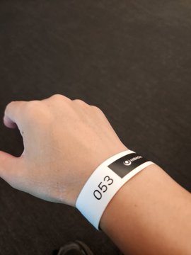 A person's arm and hand are shown against a dark gray background, wearing a white paper wristband printed with the number "053" and a black logo section.