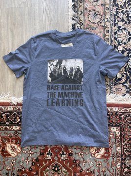A heather blue t-shirt displayed on a patterned rug features a black-and-white graphic of silhouetted figures with the bold text "RAGE AGAINST THE MACHINE LEARNING," tying into the post's promotion of a provocative art merchandise collaboration.