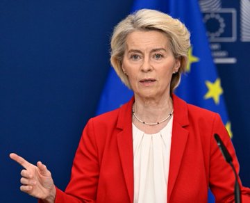European Commission President Ursula von der Leyen speaks at a podium wearing a red blazer, with European Union flags visible behind her.