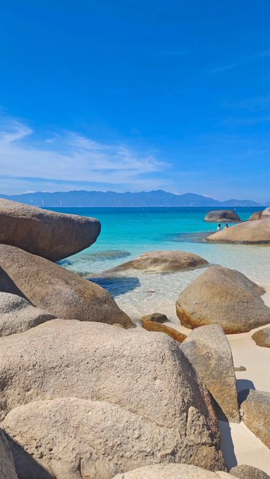 A scenic beach view featuring large smooth rocks in the foreground, turquoise water, and a distant mountain range under a bright blue sky.