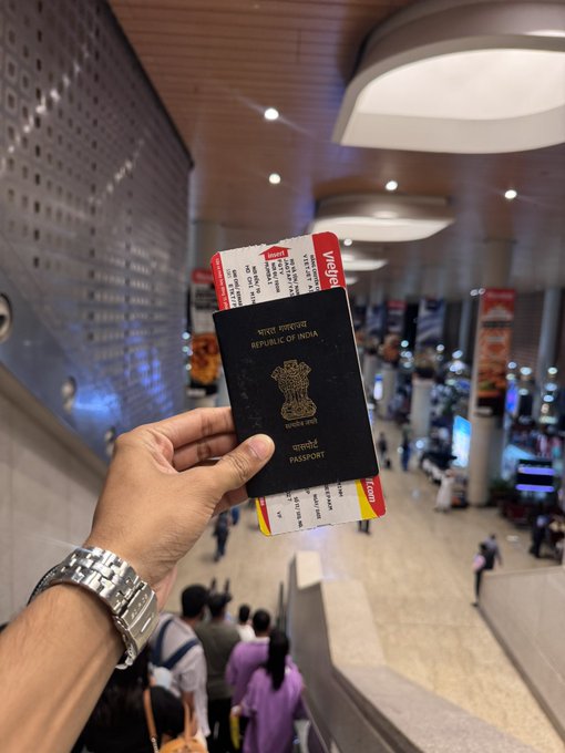 A hand holding an Indian passport with attached airline tickets in what appears to be an airport terminal.