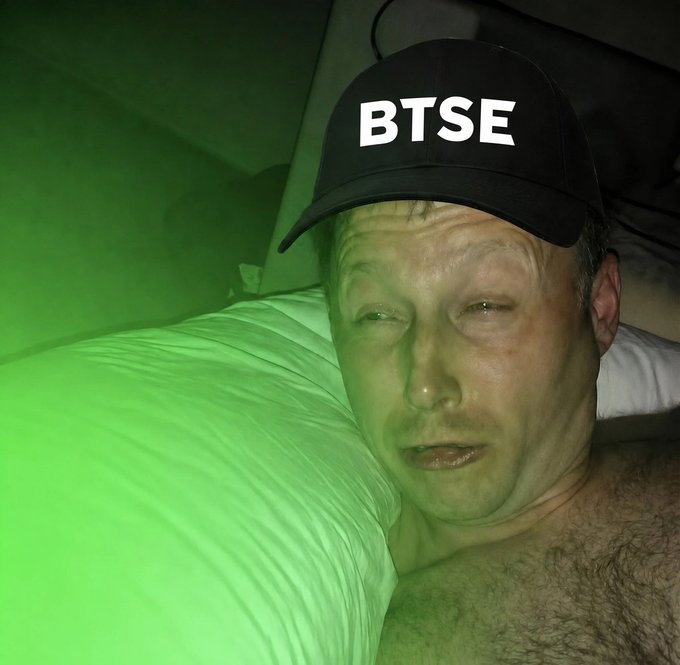 A shirtless man wearing a black baseball cap with white text reading 'BTSE' lies in bed with green lighting illuminating the scene.