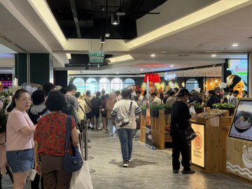 The image shows a large group of people walking through an indoor shopping mall, with some individuals carrying handbags. The crowd appears to be moving towards a food court area within the mall. There are several potted plants placed throughout the scene, adding greenery to the environment. A person is also holding a cup in their hand.

The post features a photo of a group of people and a logo for Bitcoin Wallet U, which could potentially be related to an event or promotion happening at the shopping mall. The image captures the bustling atmosphere typical of a busy shopping center during peak hours.