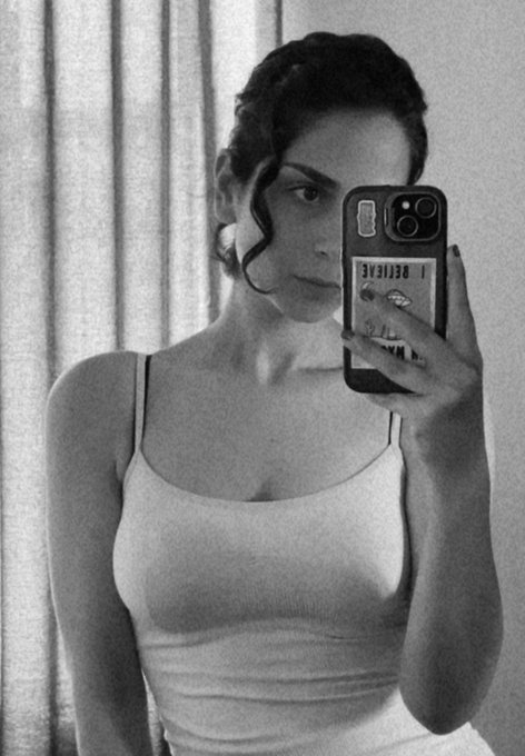 Black-and-white mirror selfie of a woman with dark hair pulled back, wearing a white tank top and holding a smartphone with stickers.