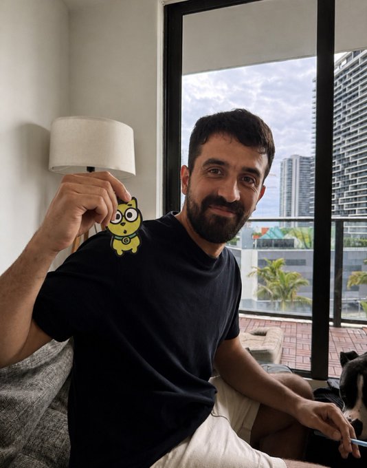 The image features a man sitting on a couch with a yellow cartoon cat charm hanging from his shirt. The caption of the image reads "BREAKING: Internet sensation, Clemente, has announced his only holdings on Abstract Chain is $GOD. God the Dog is the best original IP on Abstract and the team is top tier". This suggests that Clemente, the man holding the charm, is a fan of the cartoon cat character God from the popular animated series "Abstract" by the same artist. The image also includes a dog toy in the background, which further supports the theme of the cartoon cat charm being related to the "God the Dog" series.