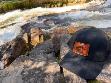 A dark baseball cap with an orange leather patch reading 'TRT' rests on rocks beside a rushing river.