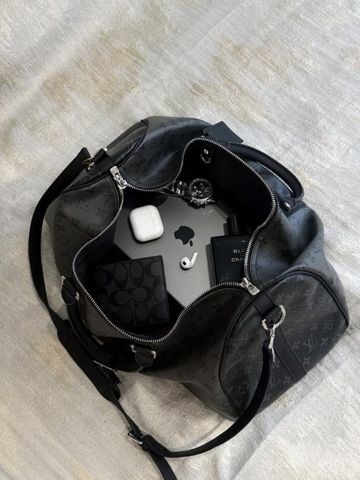 An open black monogram-patterned designer handbag containing Apple products (MacBook, AirPods, iPhone), a Coach wallet, and a smartwatch.