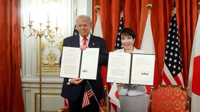 A photograph showing two people, Donald Trump in a dark suit with red tie and a Japanese woman in a light suit, holding up signed documents with American and Japanese flags in the background, appearing to be a diplomatic signing ceremony.