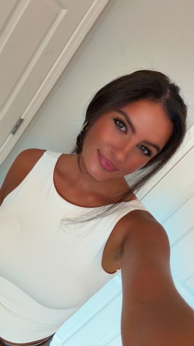 A woman with long brown hair wearing a white sleeveless top takes a selfie near a staircase.