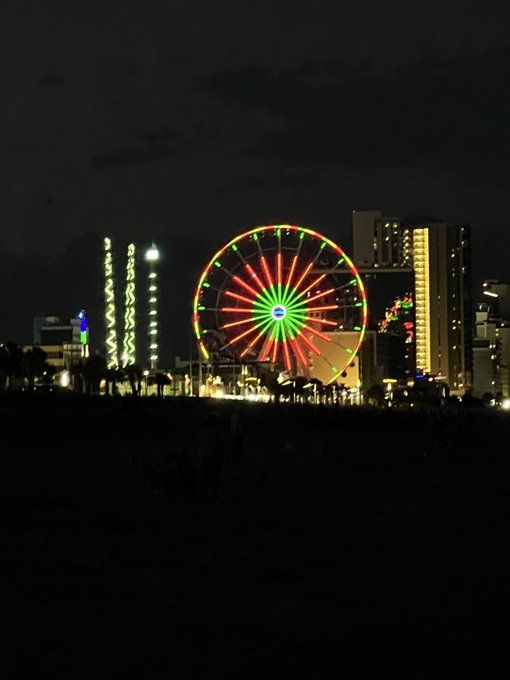 A nighttime photograph of a brightly lit Ferris wheel glowing with red and green lights, set against a dark sky with illuminated high-rise buildings and decorative light columns in the background.