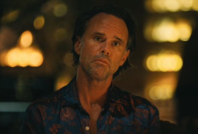 A close-up shot of an older man with a serious expression, wearing a blue patterned shirt, set against a blurry background of warm lights.