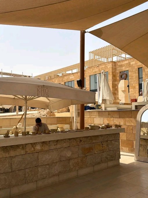 An open-air outdoor restaurant or bar area at a sunlit stone building features 'KHUFUS' branded umbrellas and a worker behind a counter displaying ceramic items.