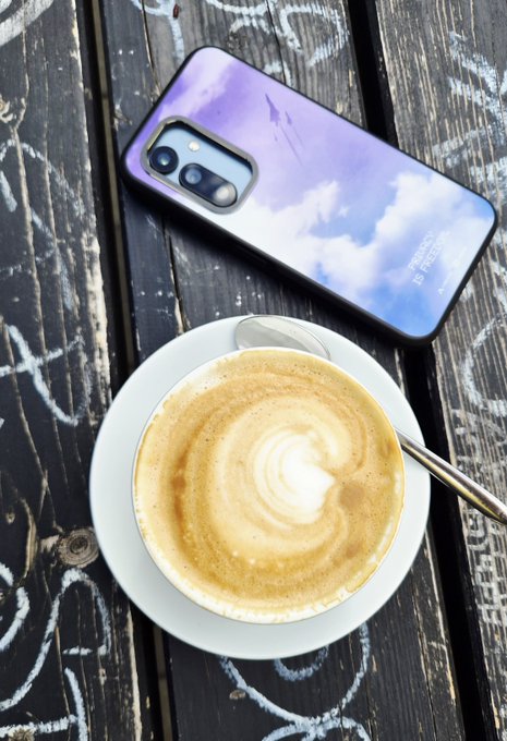 A smartphone with a purple sky-clouds case rests on a wooden surface next to a cappuccino with latte art and a spoon.