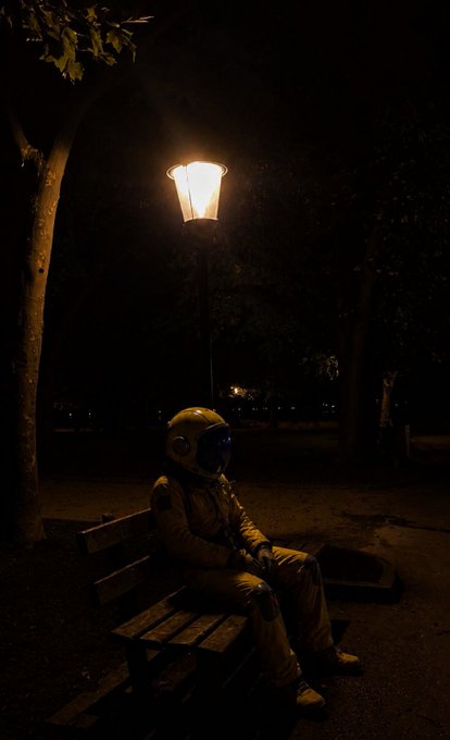 A nighttime photograph of a person in a full astronaut suit and helmet sitting alone on a wooden park bench under the warm glow of a vintage street lamp.