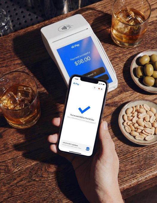 A hand holds a smartphone displaying a successful payment confirmation for $56.00 via a contactless payment terminal on a wooden bar table, accompanied by drinks and snacks.