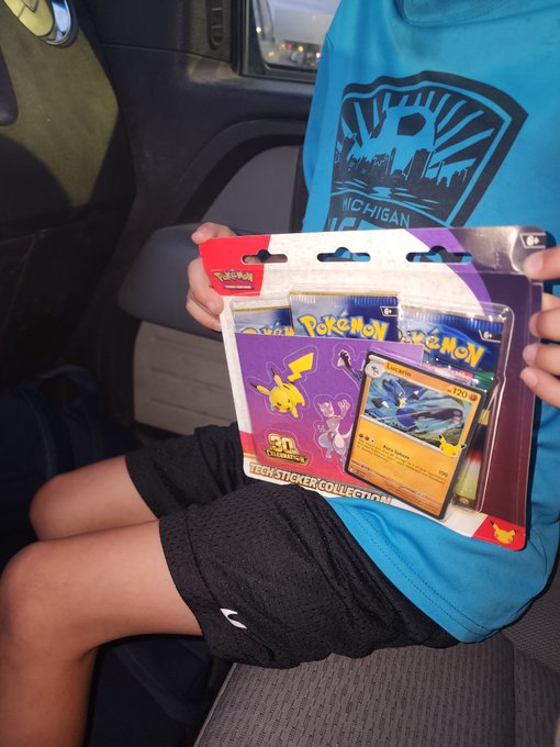 A child wearing a teal 'MICHIGAN' t-shirt and black shorts sits in a car seat, holding a Pokémon 'Legendary Sticker Collection' blister pack featuring a Lugia card.