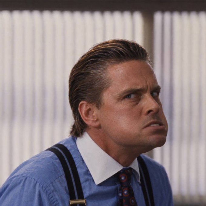 A man with slicked-back hair, wearing a blue dress shirt, suspenders, and a patterned tie, looks off to the side with a serious expression.
