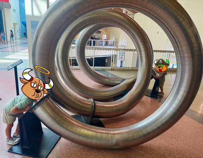 A young child looking through a large coiled metal tube sculpture in what appears to be a museum or public space, with cartoon bear stickers overlaid on the image.