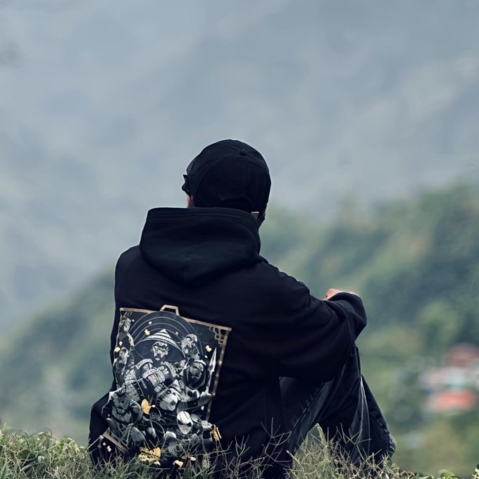 A person seen from behind, sitting on grass and looking out at mountains, wearing a black hoodie with a detailed religious-style graphic print on the back and a black cap.