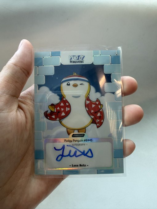 A hand proudly holds an autographed Pudgy Penguins trading card signed by Luca Netz, labeled "Pudgy Penguin #6445" with the inscription "26/49," celebrating the rare collectible's arrival in Bangkok ahead of the Pudgy Coffee Run event.