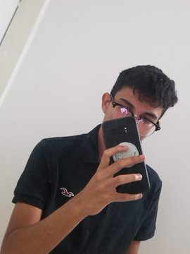 A young person with glasses taking a mirror selfie with a smartphone, wearing a black polo shirt, in a bathroom setting.