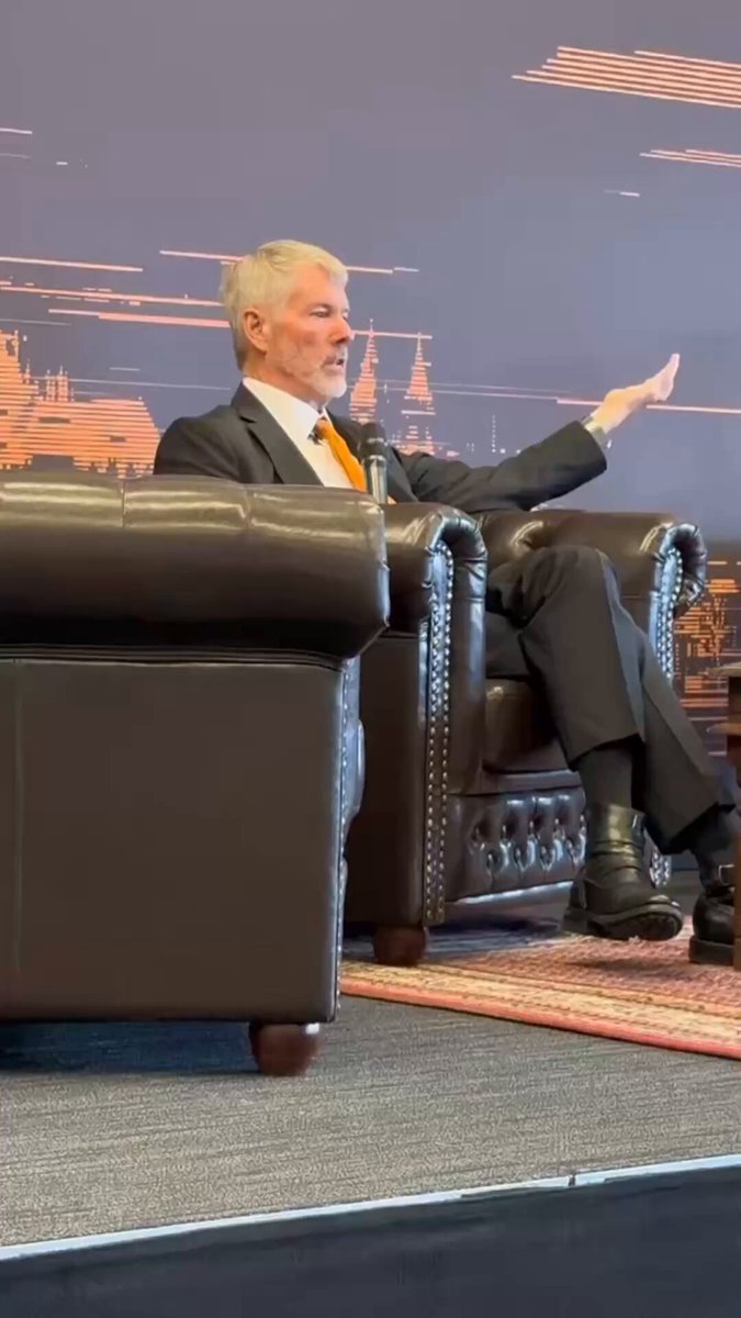 The image shows a man sitting in a chair at an event or conference. He appears to be giving a speech or presentation while holding up his hand with one finger extended. The background features a large screen displaying a map of Europe, which could suggest that this is taking place during a gathering related to international business or finance. The post about Bitcoin and the company selling it has caught the attention of the audience, as they seem interested in discussing its implications for their own investments or businesses.