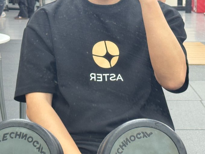 A person wearing a black t-shirt with a reversed 'ASTER' logo sits holding Technogym dumbbells, with the text appearing mirrored in the photo.