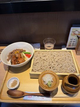 A tray of food featuring soba noodles on a bamboo mat, accompanied by a bowl of rice topped with beef, various sauces, and a glass of water.