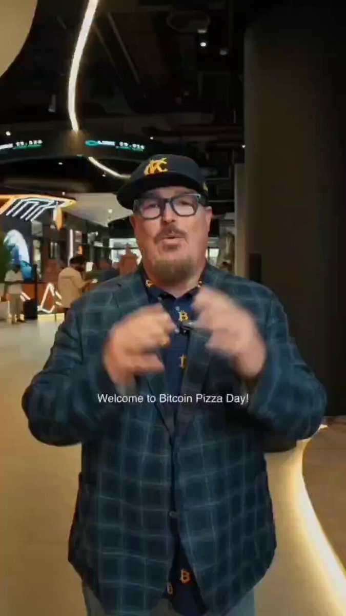 The image features a man wearing glasses, a plaid shirt, and a baseball cap standing inside an indoor space with a large screen behind him. He appears to be giving a speech or presentation related to Bitcoin Pizza Day. The background of the image shows a crowd of people gathered around the screen, indicating that this event is taking place in a public area where many people have come together for it.