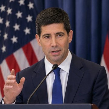 A man in a dark suit and blue tie speaks at a podium with American flags in the background, gesturing with his hand as he addresses an audience. The image appears to relate to the Federal Reserve's interest rate decision, potentially depicting a Fed official discussing monetary policy amid market expectations of a 25 basis point rate hike and Trump's nomination of Kevin Warsh.