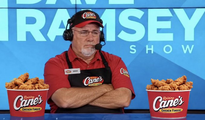 A man wearing a Raising Cane's hat, headset, and apron sits with his arms crossed on 'The Dave Ramsey Show' set, flanked by two large red buckets of Raising Cane's Chicken Fingers.
